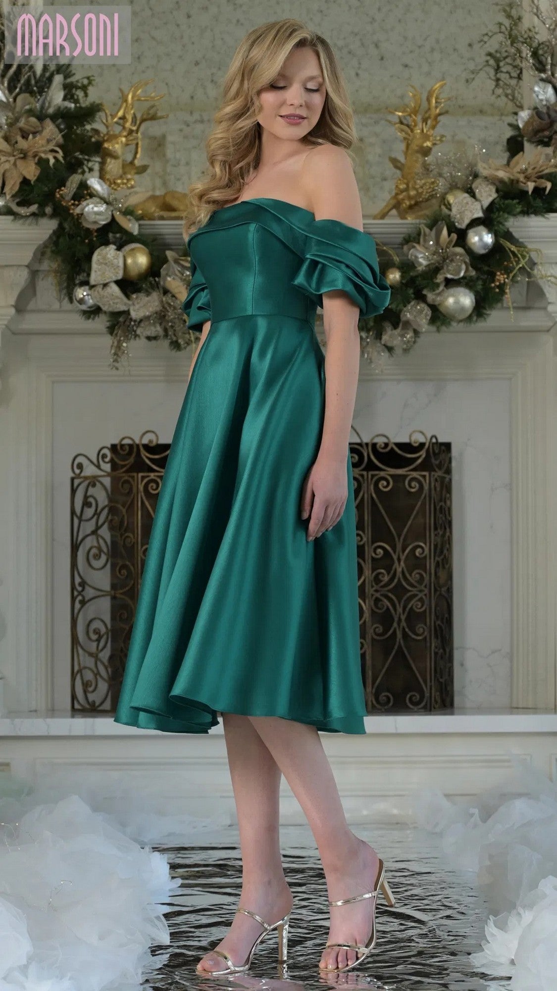 A woman in an emerald Marsoni MV1369 knee-length semi-formal dress stands indoors before a decorated white fireplace, wearing silver heels.